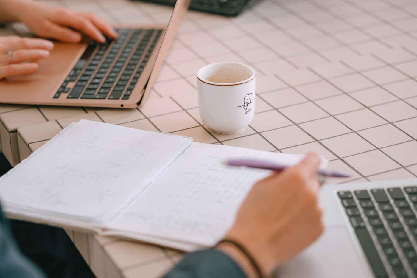 Two people planning at a table with laptops, an open notebook and a cup of coffee